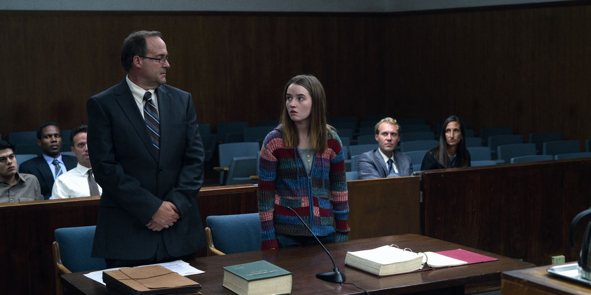 A young woman in a multicolored sweater stands in a courtroom beside a man in a navy blue suit.