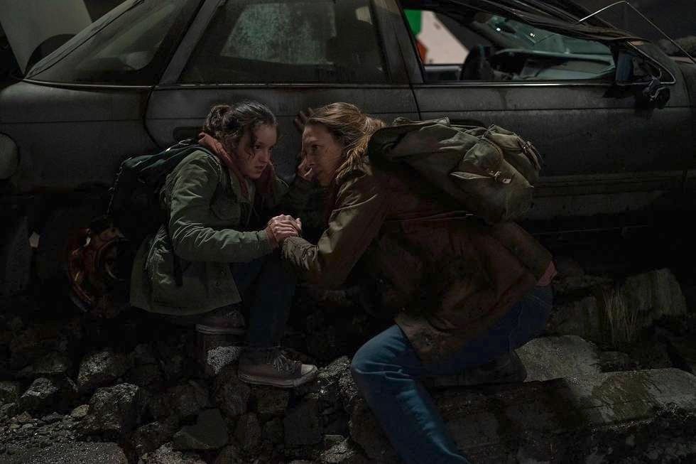 Ellie (Bella Ramsey) and Tess (Anna Torv) crouching behind a decrepit car in a still from The Last of Us