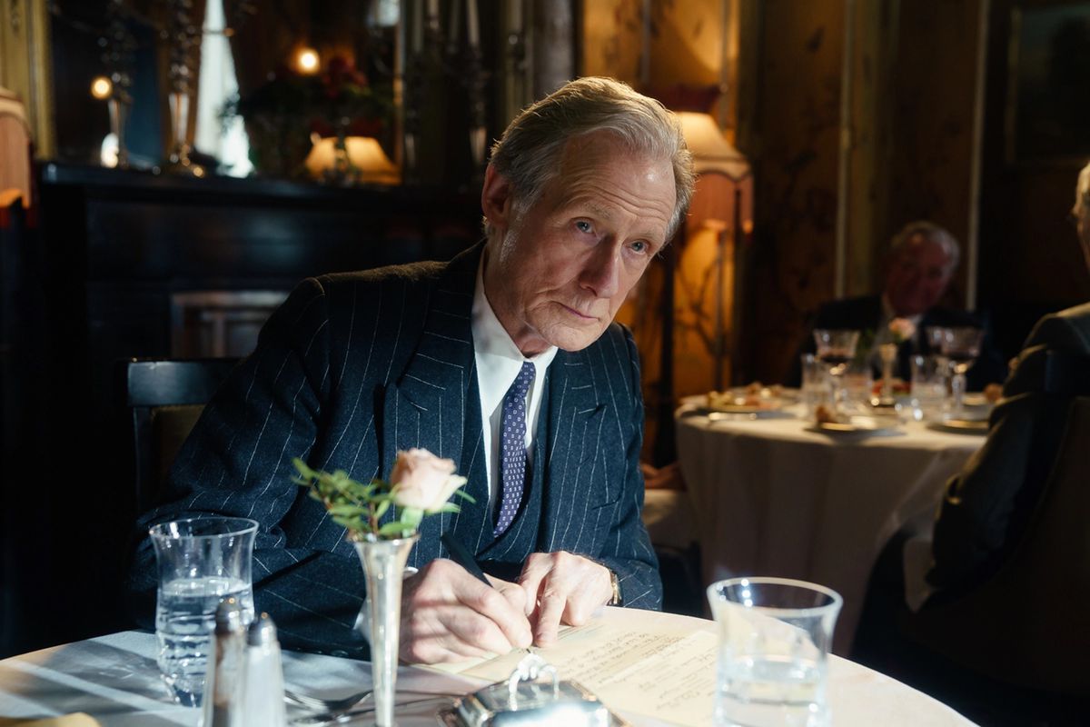 Bill Nighy, wearing a pinstripe suit, writes something at a restaurant table in Living.