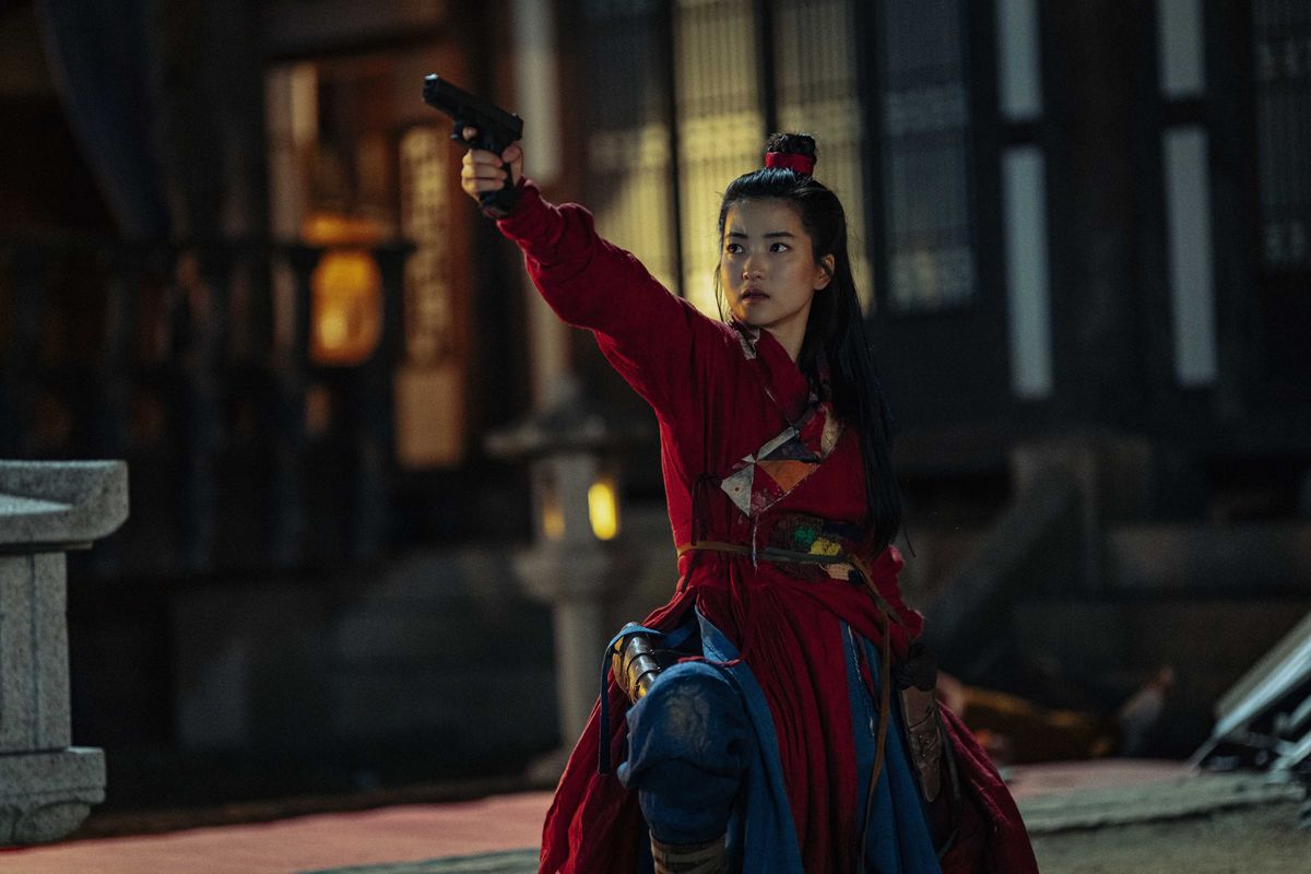 A woman (Kim Tae-ri) with her hair in a bun dressed in a red and blue kimono aims a pistol offscreen while kneeling in the middle of a courtyard.