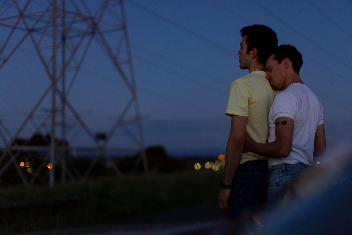 A young man (Thom Green) in a white sleeveless shirt hugs another young man (Elias Anton) in a yellow shirt from behind while staring out at a darkened sky with a powerline tower visibly in the distance.