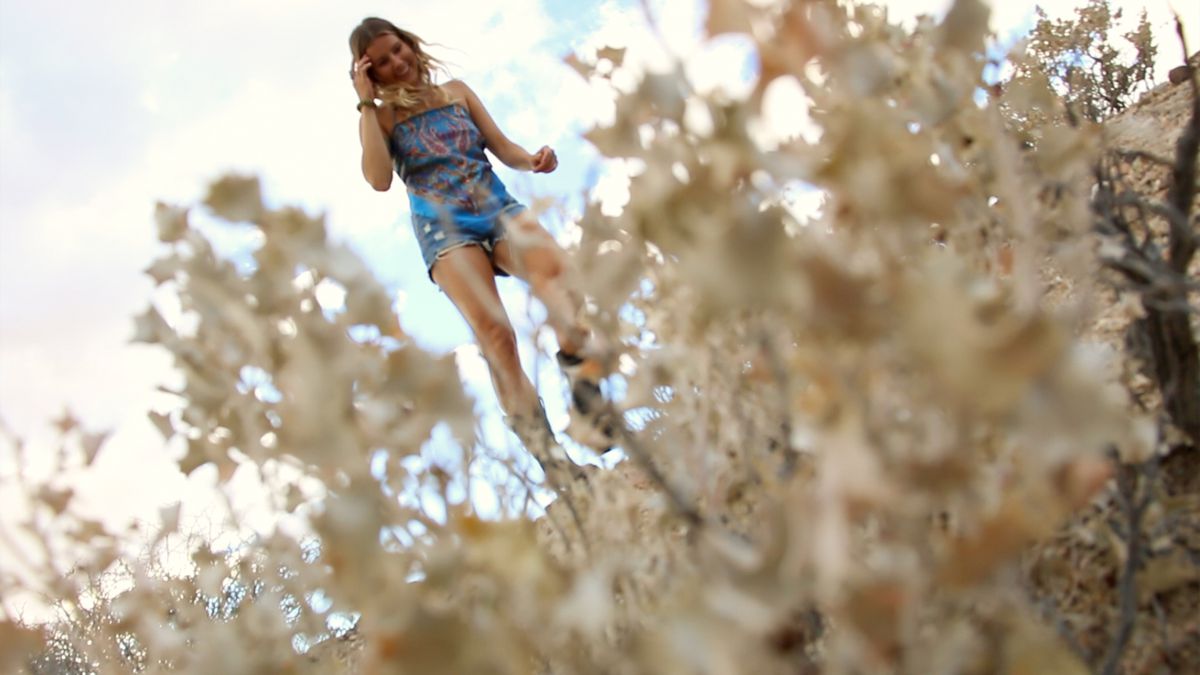 A young blonde woman wearing a colorful top iss seen through desert plants in The Outwaters.