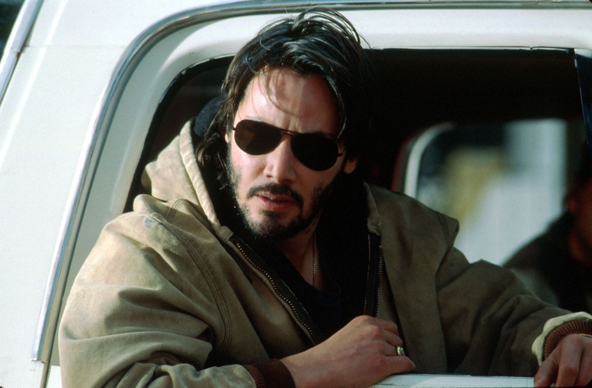 Keanu Reeves looks very cool with sunglasses, disheveled hair, and a brown jacket in the passenger seat of a car in The Gift.