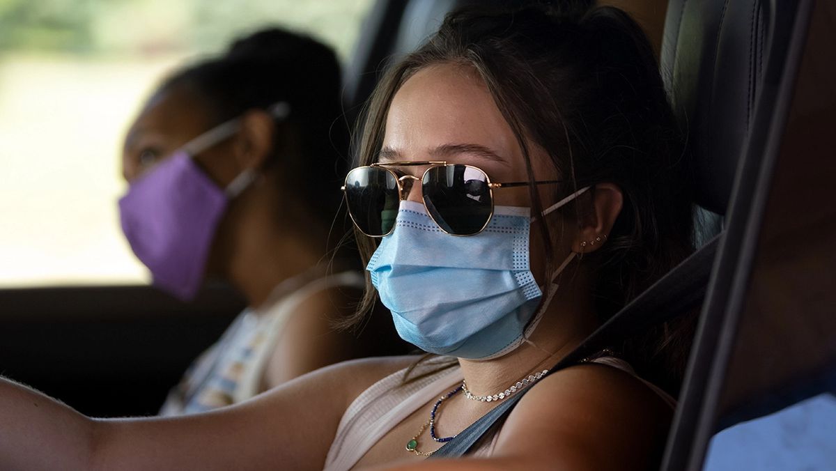 A woman wearing sunglasses and a blue face mask sits behind the wheel of a vehicle with a passenger beside them wearing a purple face mask.