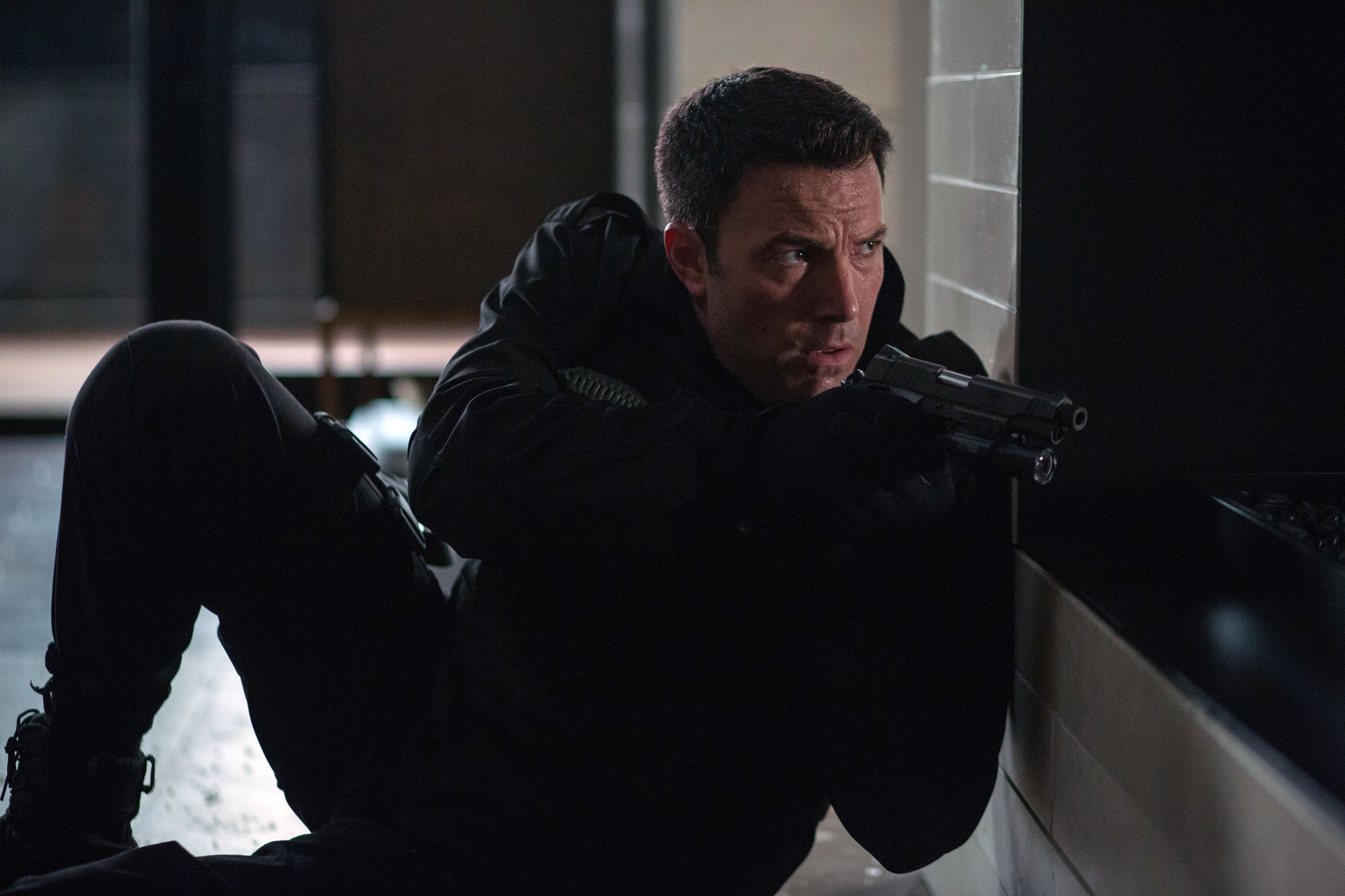 Ben Affleck with a gun wearing all black crouching behind a wall in The Accountant